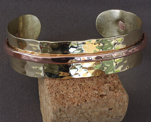 bracelets-cuivre-laiton-martele-poiconne-arthrose-mixte-clmduorivet-1.jpg Bracelet artisanal en laiton martelé et cuivre, modèle mixte riveté, posé sur un support en liège.