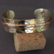 Bracelet artisanal en laiton martelé et cuivre, modèle mixte riveté, posé sur un support en liège.