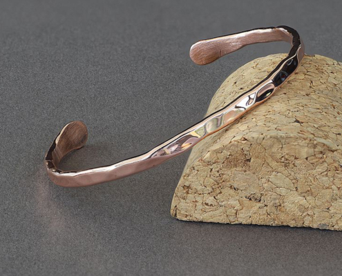 Bracelet jonc en cuivre martelé, fabriqué à la main à partir d’un fil de 3 mm, posé sur un support en liège. La surface brillante et irrégulière reflète la lumière, mettant en valeur le travail artisanal du métal.