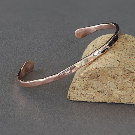 Bracelet jonc en cuivre martelé, fabriqué à la main à partir d’un fil de 3 mm, posé sur un support en liège. La surface brillante et irrégulière reflète la lumière, mettant en valeur le travail artisanal du métal.