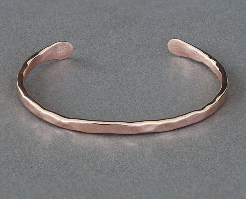 bracelet-cuivre-fil-martele-02.jpg Bracelet jonc en cuivre martelé vu de face, posé à plat sur fond gris, montrant sa forme arrondie et sa texture brillante façonnée à la main.