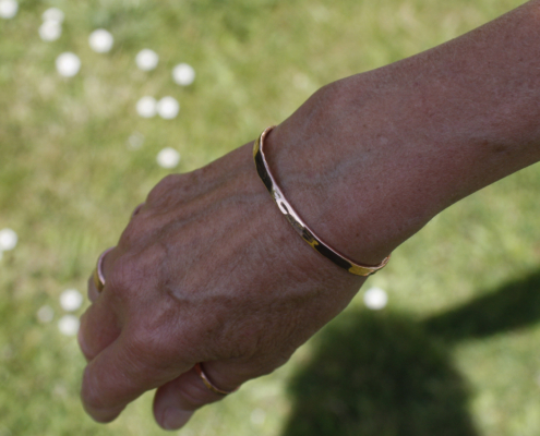bracelet-cuivre-fil-martele-porte-femme-03.jpg Bracelet jonc en cuivre martelé porté au poignet d’une femme, en extérieur sur fond d’herbe verte, mettant en valeur l’éclat du métal au soleil.