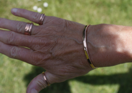 Main d’une femme portant un bracelet jonc en cuivre martelé accompagné de plusieurs bagues en cuivre, en extérieur sur fond d’herbe verte.