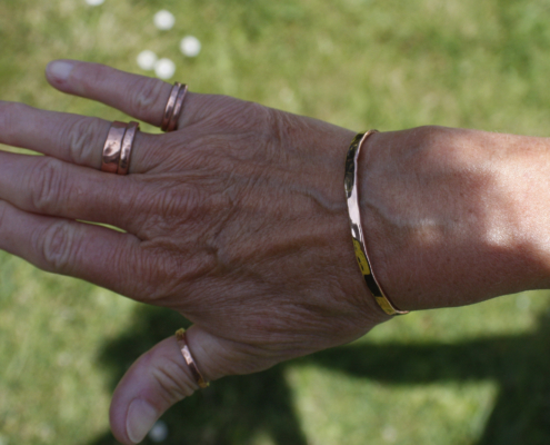 bracelet-cuivre-fil-martele-porte-femme-04.jpg Main d’une femme portant un bracelet jonc en cuivre martelé accompagné de plusieurs bagues en cuivre, en extérieur sur fond d’herbe verte.