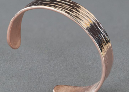 Bracelet en cuivre martelé avec finition striée intense, présenté sur fond gris, vu en perspective.