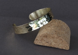 Bracelet laiton martelé chat présenté en équilibre sur un support en liège, lumière naturelle reflétée sur le métal poli.