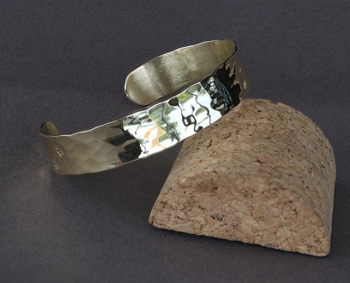 Bracelet laiton martelé chat présenté en équilibre sur un support en liège, lumière naturelle reflétée sur le métal poli.