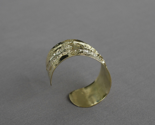 Bracelet large en laiton texturé, vu de profil, sur fond gris uni.