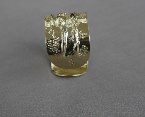 Gros plan sur l’extrémité d’un bracelet en laiton texturé, finition brute et martelée, posé sur fond gris.