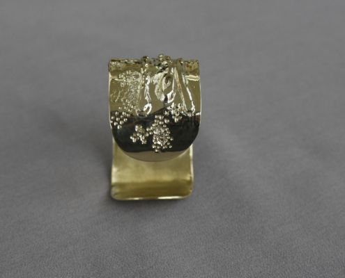 Vue frontale d’un bracelet en laiton martelé avec des textures organiques et granuleuses sur fond gris.