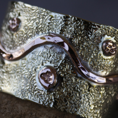 Bracelet en laiton et cuivre martelé, décoré de rivets en relief et d’une ondulation centrale brillante, sur fond de texture granuleuse