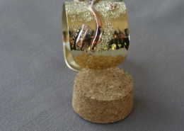 Bracelet en laiton brillant orné d’un motif ondulé en cuivre rouge sur fond texturé, présenté debout sur un bouchon de liège.
