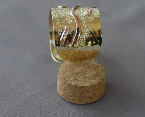 Bracelet en laiton brillant orné d’un motif ondulé en cuivre rouge sur fond texturé, présenté debout sur un bouchon de liège.